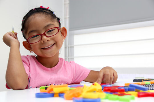 A young girl smiles for the camera while playing with puzzle piees on the table in front of her